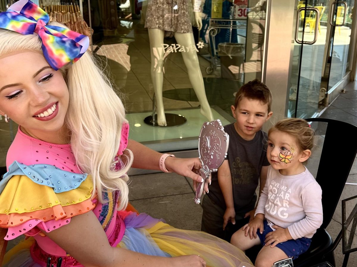 A face painter at Lucky Stars Market holds up a mirror for a little girl and her brother to see a  yellow butterfly painted on her left cheek.