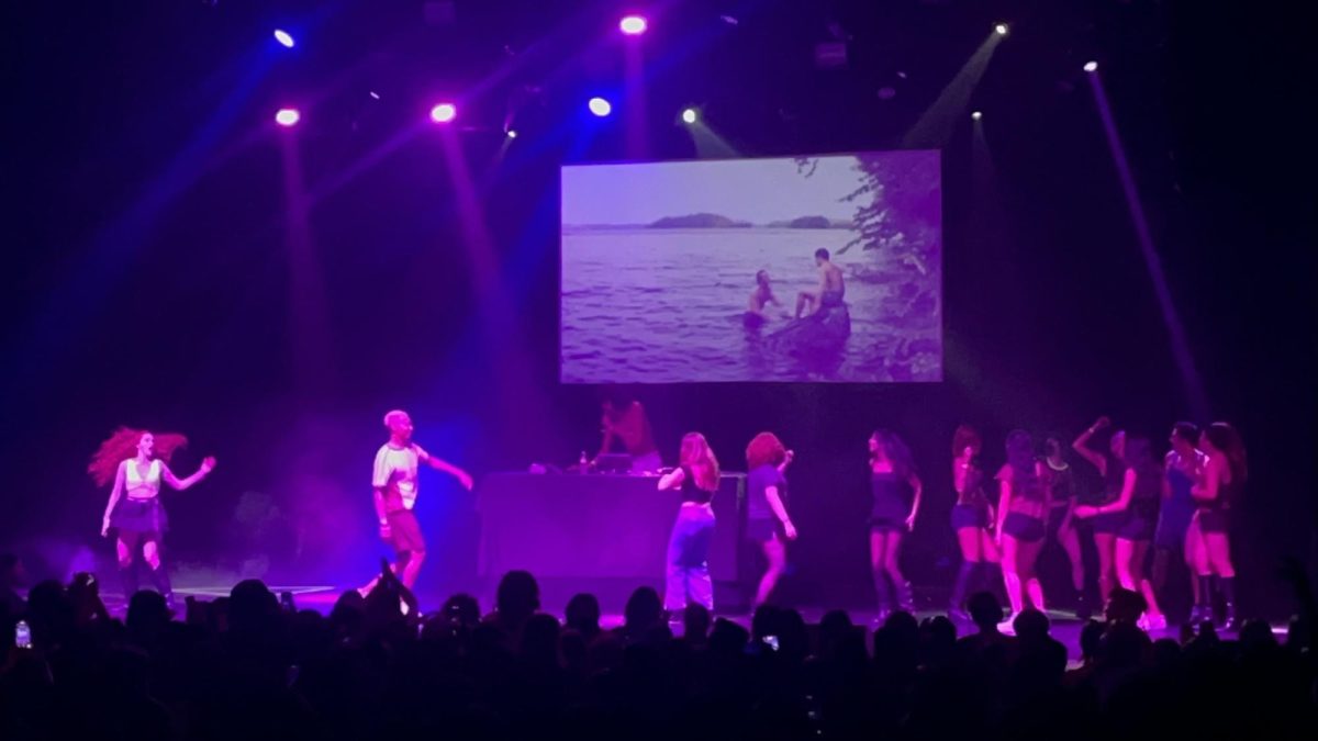Fans at the "Heated RivalRave" dance on stage as clips from "Heated Rivalry" play on the screen