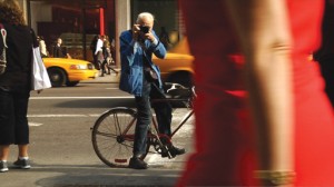 Photographer Bill Cunningham takes a shot of an unaware subject on the crowded New York City street on his bike. Bored by the world of fashion photography, Bill takes to the streets for photos of everyday people, Courtesy of First Thought Films / Zeitgeist Films